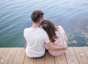 couple-sitting-wooden-pier-her-head-resting-shoulder-him
